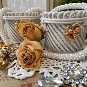 May include: Two white woven baskets with gold grape cluster accents are displayed with dried roses, pearl necklaces, and vintage jewelry. The baskets have a decorative, open weave design. The scene is set on a lace doily.