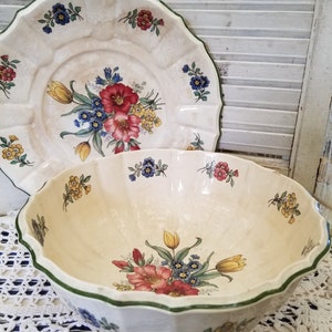 May include: Cream-colored ceramic serving bowl and plate set with a floral design. The bowl and plate have a scalloped edge with a green trim. The floral design includes red, yellow, and blue flowers.