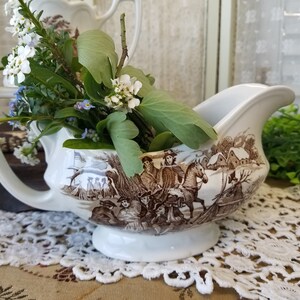May include: White porcelain gravy boat with a brown and white winter scene design. The scene features people on horseback and a small village. The gravy boat is filled with white and blue flowers.