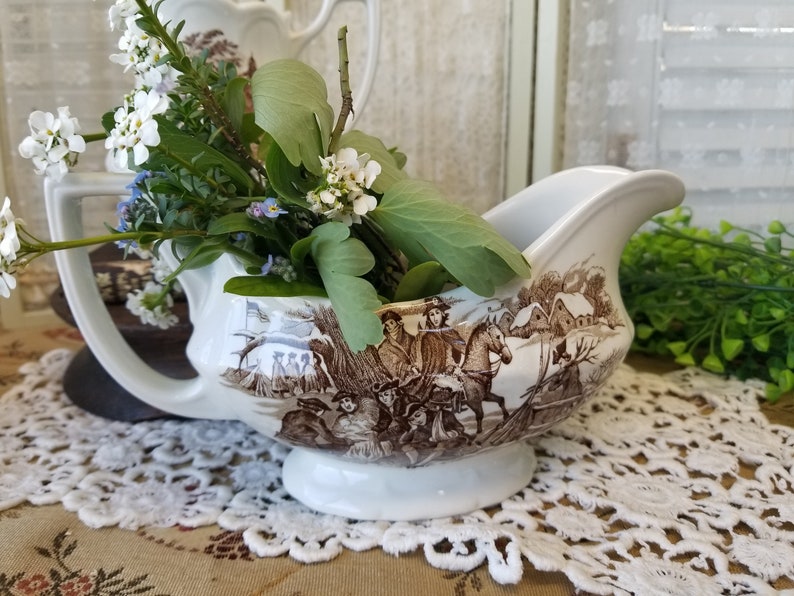 May include: White porcelain gravy boat with a brown and white winter scene design. The scene features people on horseback and a small village. The gravy boat is filled with white and blue flowers.