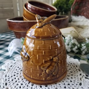 May include: A brown ceramic honey pot shaped like a beehive, with a lid featuring a bee. The pot has a textured surface and embossed bee details. Other brown ceramic bowls are in the background. The honey pot is on a white crocheted doily.