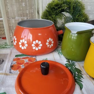 May include: An orange enamelware pot with white daisy designs and a matching lid with a black knob. A green and yellow enamelware pot are also visible. The pots are on a patterned tablecloth.