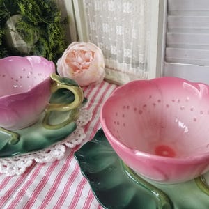 May include: Two pink ceramic teacups shaped like flowers, with matching green leaf-shaped saucers. The teacups have small holes and are set on a red and white striped tablecloth. A decorative wreath and a flower are in the background.