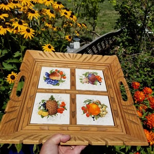 May include: A wooden serving tray with four square tile inserts, each depicting a different arrangement of fruits. The tray has carved edges and integrated handles. The fruits include grapes, peaches, plums, a pineapple, and an orange.