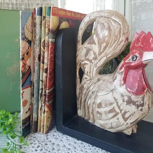May include: A rooster-shaped bookend with a red comb and wattle, and a distressed white and brown finish. The bookend holds a stack of vintage cookbooks and a green book. The text "173 Recipes from the South" is visible on one book.