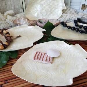 May include: Several off-white, fish-shaped decorative plates with a textured surface. Some plates hold decorative items like a black beaded necklace, a brown and tan beaded necklace, a small woven item, and a white shell. The plates are arranged on a bamboo mat.