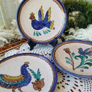 May include: Three round ceramic plates with cream-colored centers and blue rims. Each plate features hand-painted designs of birds in blue, yellow, and green, along with floral motifs. The plates are decorated with brown accents and small initials.