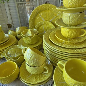 May include: A collection of bright yellow ceramic dinnerware featuring a textured leaf pattern. The set includes multiple stacked dinner plates, salad plates, bowls, and cups with saucers. Each piece displays a consistent embossed foliage design.
