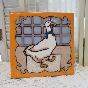 May include: Square ceramic tile with a hand-painted illustration of a white goose wearing a blue scarf. The goose is set against a window design with a yellow border. The tile is likely for decorative use.