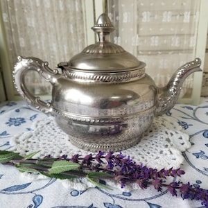 May include: A silver teapot with a decorative handle and a round body. The teapot has a shiny finish and a beaded trim around the top. The teapot is sitting on a white crocheted doily.