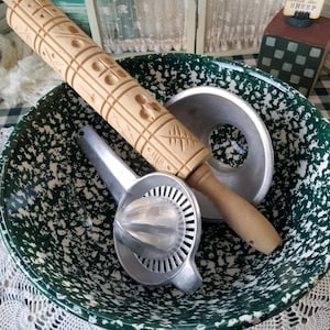 May include: A vintage kitchen scene featuring a green and white speckled bowl holding a wooden rolling pin, a metal juicer, and a metal lid. The rolling pin is light brown with decorative carvings. The bowl is on a checkered tablecloth.