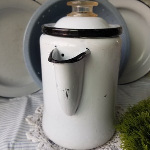 May include: White enamel coffee pot with a black rim and a black spout. The pot has a white lid with a clear plastic knob.