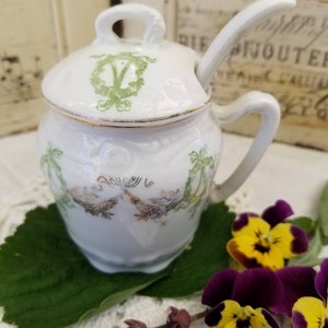 May include: White porcelain sugar bowl with a lid and a small spoon. The bowl has a green and gold floral design. The bowl is sitting on a white lace tablecloth with green leaves and purple flowers.