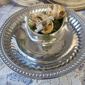 Puede incluir: Una bandeja plateada con una zona central para salsas, con huevos de Pascua decorados en musgo verde. La bandeja tiene un borde con cuentas y una superficie brillante, ideal para aperitivos.