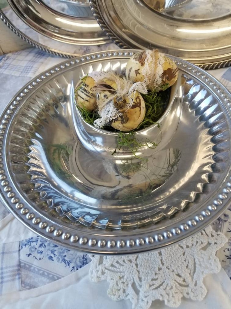 Puede incluir: Una bandeja plateada con una zona central para salsas, con huevos de Pascua decorados en musgo verde. La bandeja tiene un borde con cuentas y una superficie brillante, ideal para aperitivos.