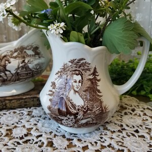 May include: A pair of vintage white ceramic creamer and sugar bowl set with brown floral and landscape designs. The creamer features a woman in a brown dress and a tree in the background. The sugar bowl features a horse and a house in the background.