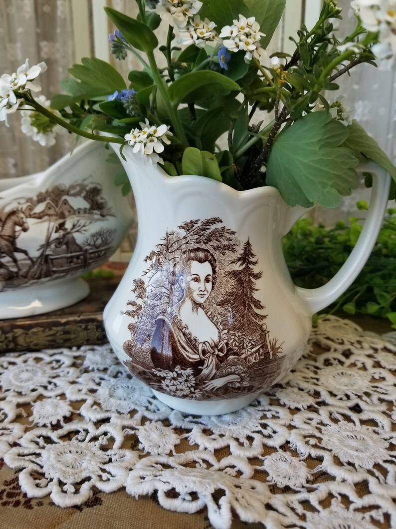 May include: A pair of vintage white ceramic creamer and sugar bowl set with brown floral and landscape designs. The creamer features a woman in a brown dress and a tree in the background. The sugar bowl features a horse and a house in the background.