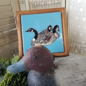 May include: A wooden duck decoy with a weathered appearance, featuring a maroon head and a blue beak. A framed tile with a painting of three geese on a blue background is in the background. The scene evokes a rustic, vintage aesthetic.