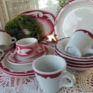 May include: A collection of white ceramic dishware with a burgundy trim. The set includes cups, saucers, bowls, and plates. The plates have a decorative burgundy design. The items are arranged on a white lace tablecloth.