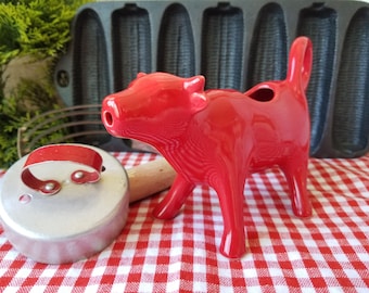 May include: A glossy red ceramic cow creamer stands on a red and white checkered tablecloth. A metal biscuit cutter and a cast iron corn-shaped baking pan are also visible. The cow creamer is a kitchenware item.