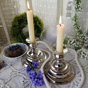 May include: Two lit ivory candles in silver candle holders. A green wreath and a small bowl with a dessert are in the background. A strand of pearls and purple flowers are arranged on a white lace tablecloth.