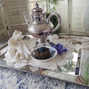 May include: A silver tea set with a decorative teapot and a serving tray. The tray holds a small cake, white gloves, a pearl necklace, and a small blue and white dish. The scene is set on a blue and white patterned surface.