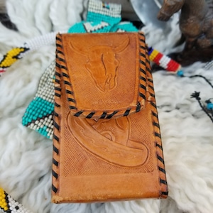 May include: A tan leather case with a flap closure, featuring embossed designs of a bull skull and a sombrero. The edges are stitched with black thread. The case is rectangular and appears to be a phone case or small wallet.