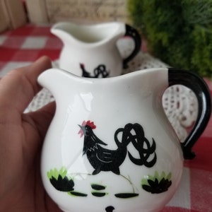 May include: Two white ceramic creamer pitchers with black handles. Both pitchers feature a black rooster design with green accents. One rooster has a red comb.