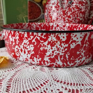 May include: A vintage enamelware roasting pan with a red and white speckled pattern. The pan has a black rim and handles. The lid is visible in the background. The pan is sitting on a white lace tablecloth.