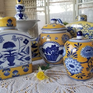 May include: A collection of decorative ceramic jars and containers. The pieces feature a combination of yellow, white, and blue colors. The designs include floral patterns and ornate details. The items are arranged on a white lace tablecloth.