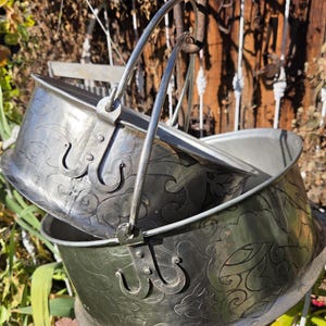 May include: Two stacked silver-toned metal buckets with swing handles. Each bucket features a repeating embossed scroll and leaf pattern on the exterior surface and a metal bracket attachment for the handle on each side.