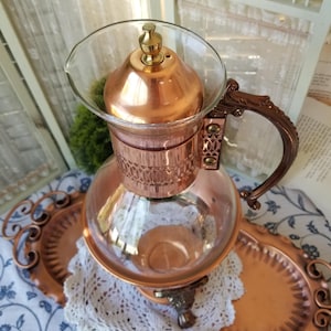 May include: A copper-colored coffee maker with a glass top and handle. The coffee maker sits on a copper-colored tray with a decorative edge. A white lace doily is beneath the coffee maker.