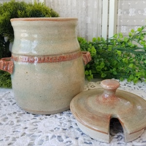 May include: A ceramic jar with a lid, both glazed in a light green and brown color. The jar has a rounded body and a wide mouth, with a brown band around the middle. The lid has a small spout on one side.