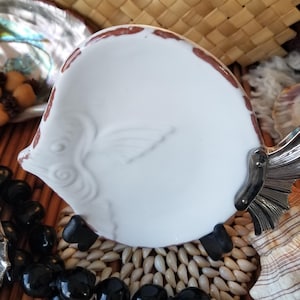 May include: A white ceramic fish-shaped dish with brown trim and a silver tail fin. The dish is surrounded by a black beaded necklace, seashells, and a woven mat. The background includes a woven basket.