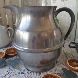 May include: A silver-toned metal pitcher with a rounded, bulbous body, a wide pouring spout, and a curved handle. The pitcher features a smooth, polished finish with a distinct horizontal ridge encircling the center of the vessel's exterior.