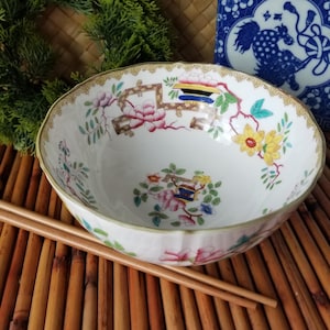 May include: A white porcelain bowl with a floral design featuring pink, yellow, and blue flowers. The bowl has a scalloped edge and is accompanied by wooden chopsticks. A blue and white decorative box is in the background.