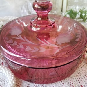 May include: A round, translucent pink glass candy dish with a matching lid. The lid features a central knob handle and is decorated with etched floral patterns, including leaves and blossoms. The base of the dish has a smooth, circular design.