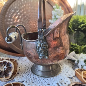 May include: A copper coal scuttle featuring a curved handle with a white and blue ceramic grip, a wide pouring spout, and a pedestal base. The sides of the vessel are adorned with metal lion head accents where the handle attaches to the body.