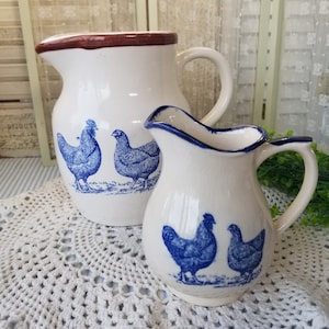 May include: Two ceramic pitchers with blue and white chicken designs. The larger pitcher has a brown rim and a handle. The smaller pitcher has a blue rim and a handle.