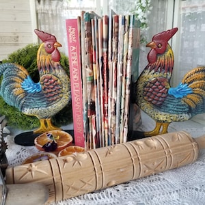 May include: Two colorful rooster bookends flank a stack of books and a rolling pin. The rooster bookends are painted with yellow, blue, and red hues. The rolling pin is made of wood and has a decorative pattern.