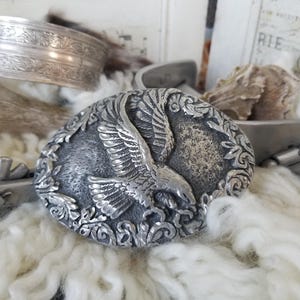 May include: Silver belt buckle with an eagle design in flight, surrounded by ornate floral details.