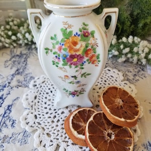 May include: A white porcelain vase with two handles, featuring a colorful floral pattern of orange, pink, and purple flowers with green leaves. The rim and handles are accented with a thin gold trim, and the vase has a flared base.