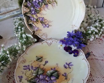 Pode incluir: Dois pratos antigos de porcelana com bordas onduladas. Cada prato apresenta um desenho pintado à mão de violetas roxas e folhas verdes sobre um fundo creme. As bordas são acentuadas com acabamentos dourados, adicionando um toque de elegância.