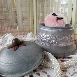 May include: A silver metal powder puff container with a floral design and a pink powder puff. The container has a wooden knob on the lid.