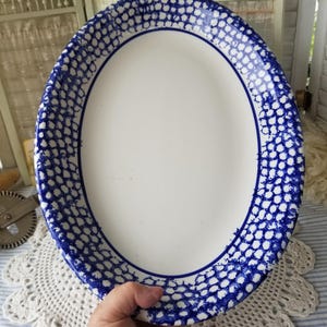 May include: An oval-shaped white platter with a blue dotted border. The rim has a dark blue line. The platter is held by a person wearing a red sweater. The background includes a white lace doily and a striped fabric.