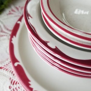 May include: A stack of white ceramic plates and bowls with a red border. The plates have a decorative red design around the edges. The bowls and plates are stacked on top of each other.