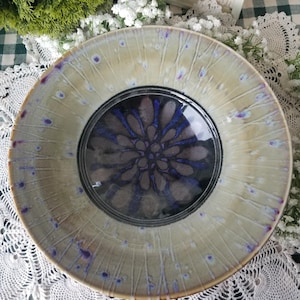 May include: A round ceramic bowl featuring a wide, light beige rim with radiating streaks and small purple-blue spots. The deep center of the bowl displays a dark, circular pattern with a vibrant blue, petal-like floral design at its core.