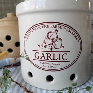 May include: A white ceramic garlic keeper with a maroon graphic and text. The graphic features garlic bulbs and the text reads "FRESH FROM THE FARMERS MARKET" and "GARLIC California Grown Since 1972". The keeper has several small holes.