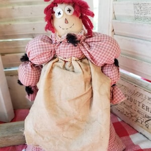 May include: A handmade rag doll with a fabric body and red yarn hair. The doll has button eyes, a stitched smile, and a red and white checkered dress. The doll's apron is a light tan color. The doll is sitting on a red and white checkered surface.