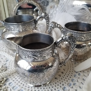 May include: Three silver creamer pitchers with ornate floral designs. The pitchers have a rounded body and a handle with a decorative finial. The pitchers are sitting on a white lace tablecloth.